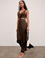 model wearing a brown dress with cut-out design, holding a white bag on a plain background.