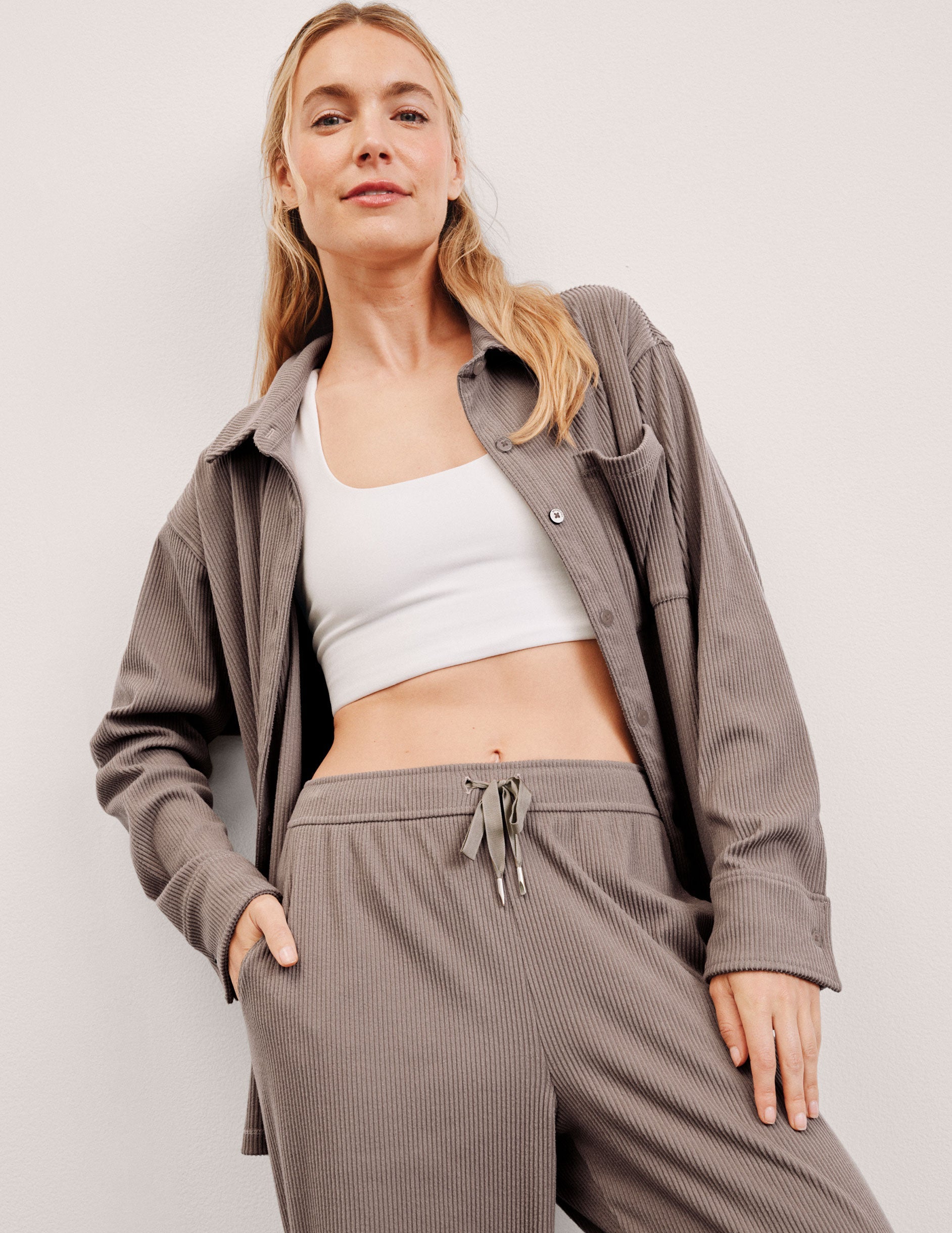 Woman wearing a brown corduroy button up shirt, white crop top, and brown corduroy pants against a plain background. 