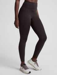 person wearing brown high-waisted long leggings with pockets on a plain background.