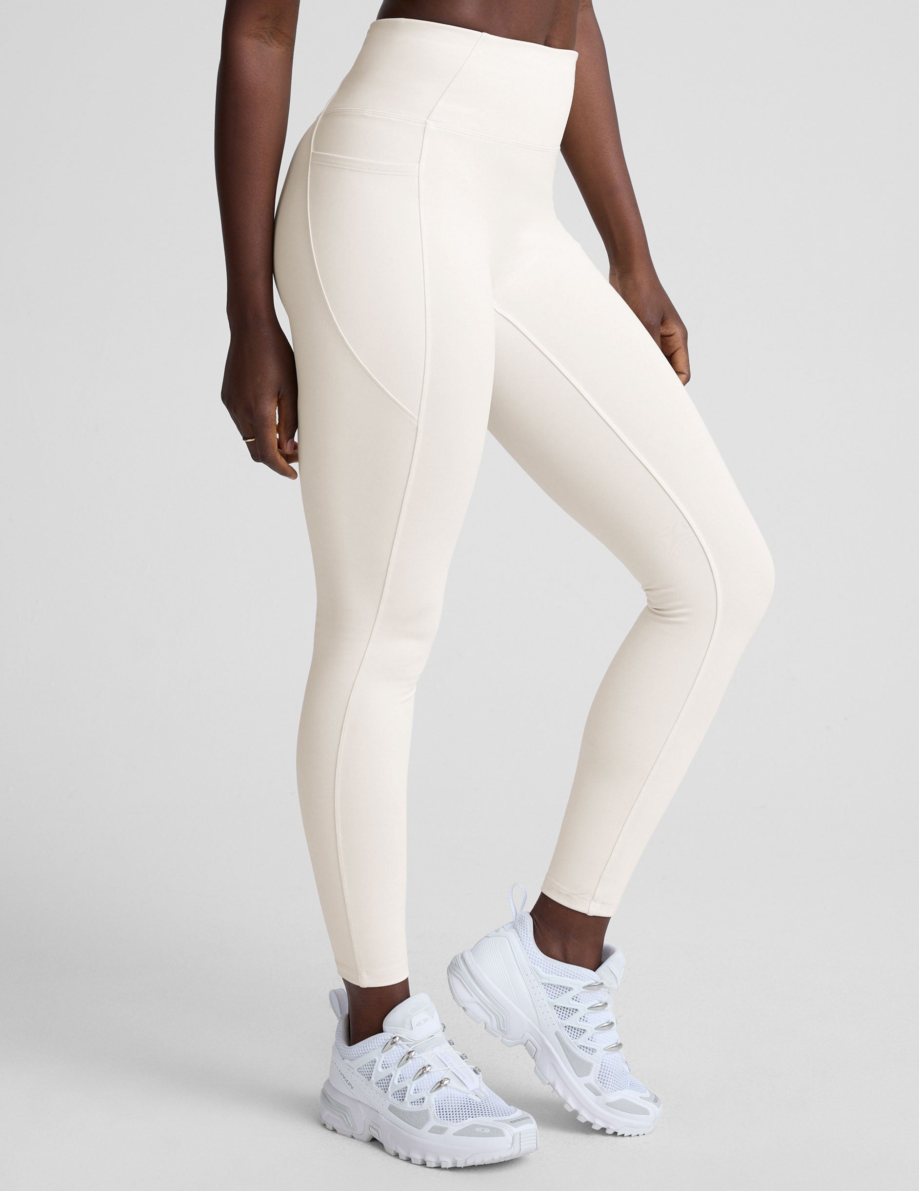 woman wearing white high-waisted long length leggings with pockets on a plain background.