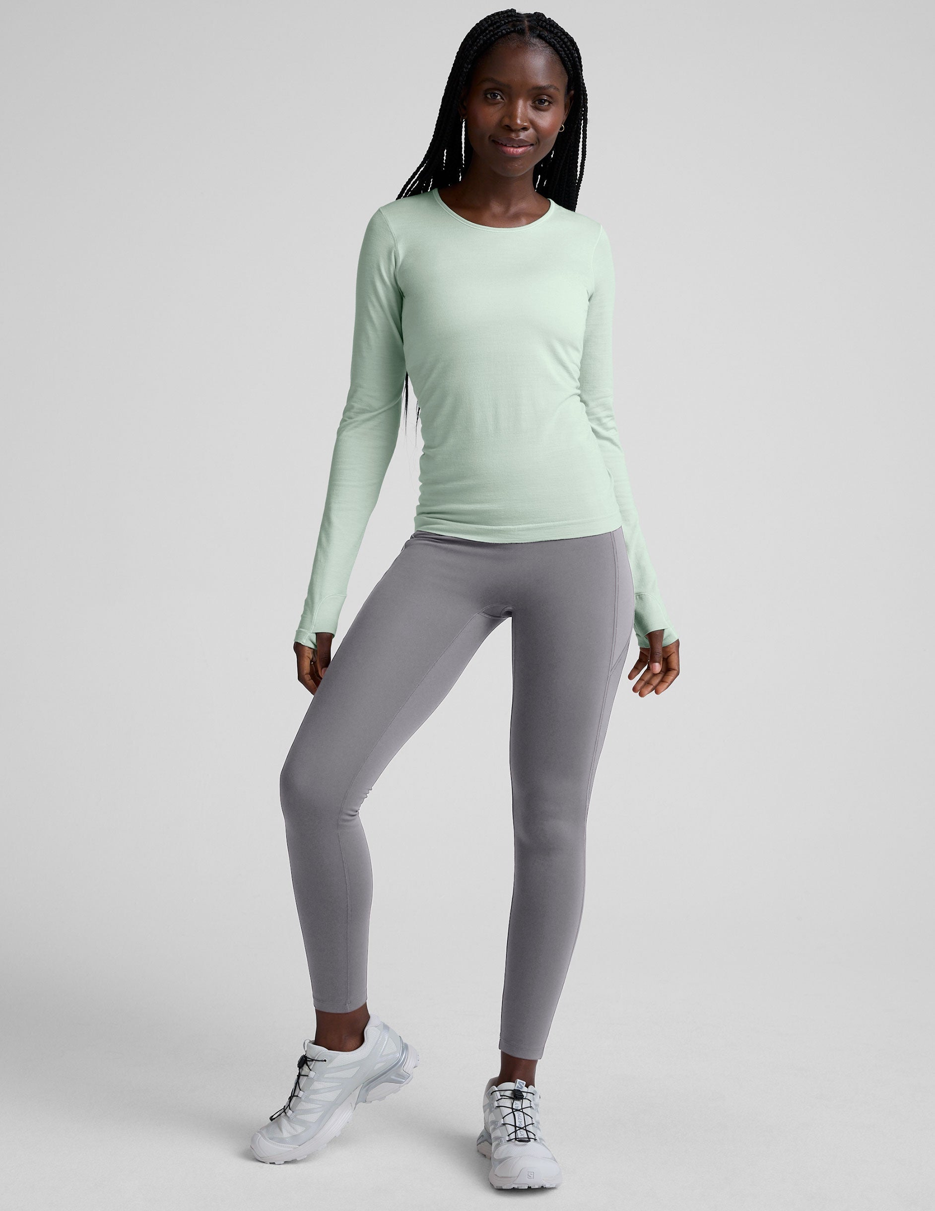 woman wearing a green long-sleeve top and gray high-waisted long length leggings on a plain background.