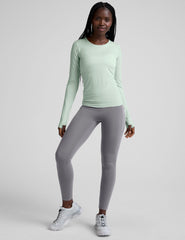 woman wearing a green long-sleeve top and gray high-waisted long length leggings on a plain background.
