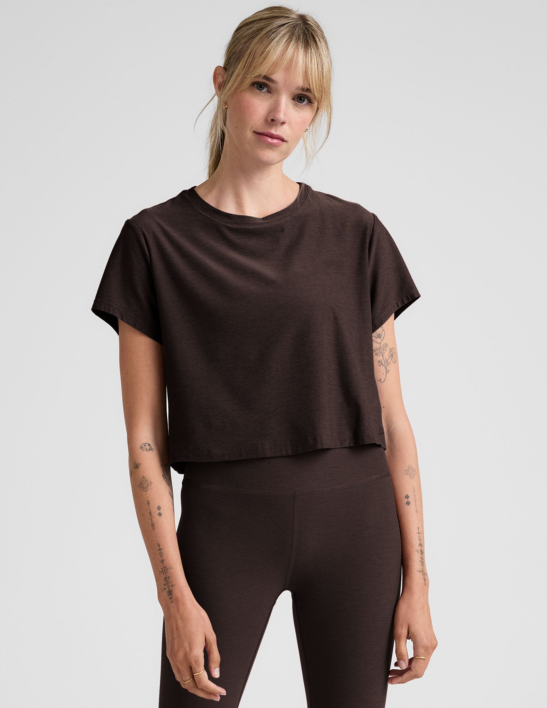 woman wearing a brown cropped t-shirt and leggings on a plain background.