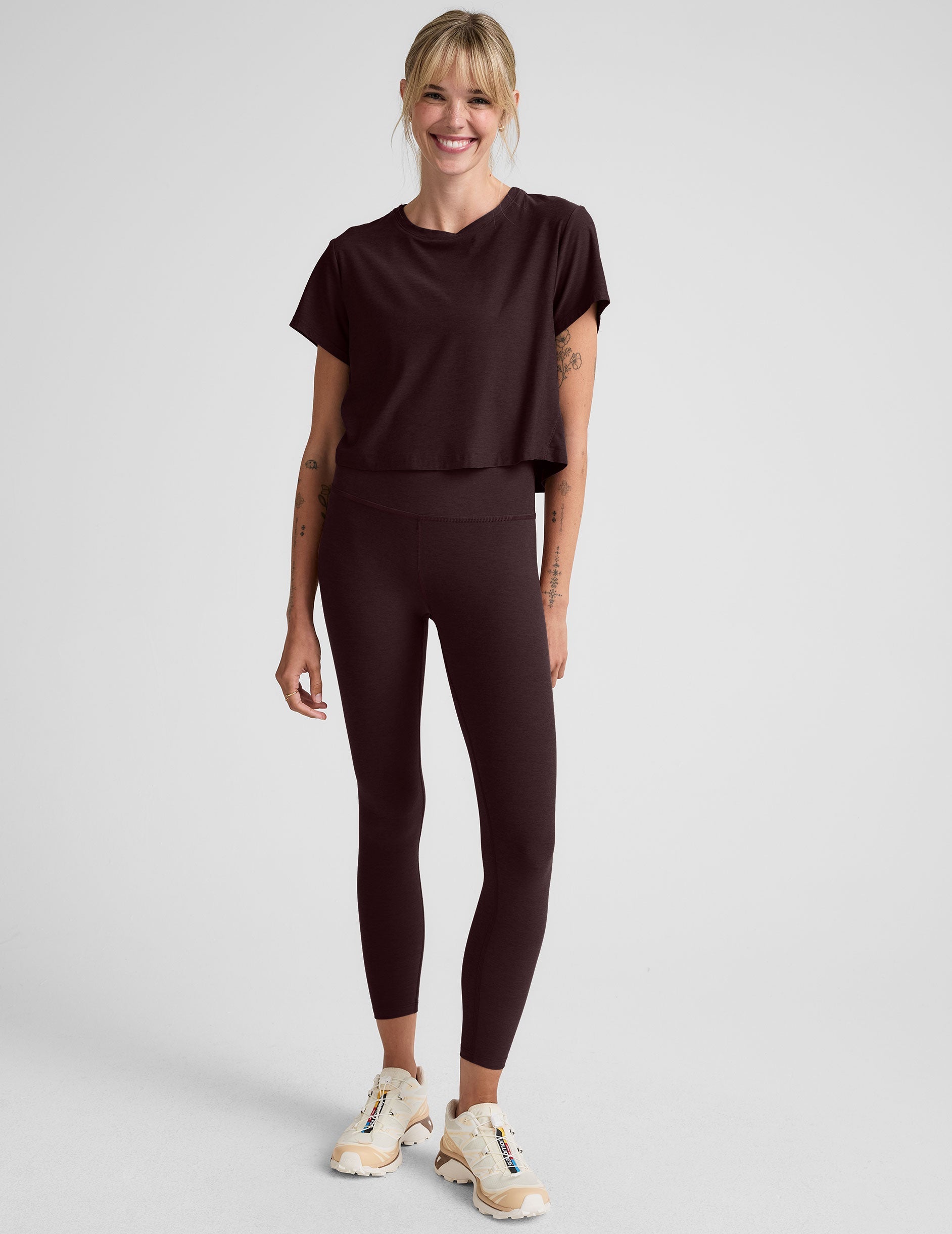 woman wearing a brown cropped t-shirt and leggings on a plain background.