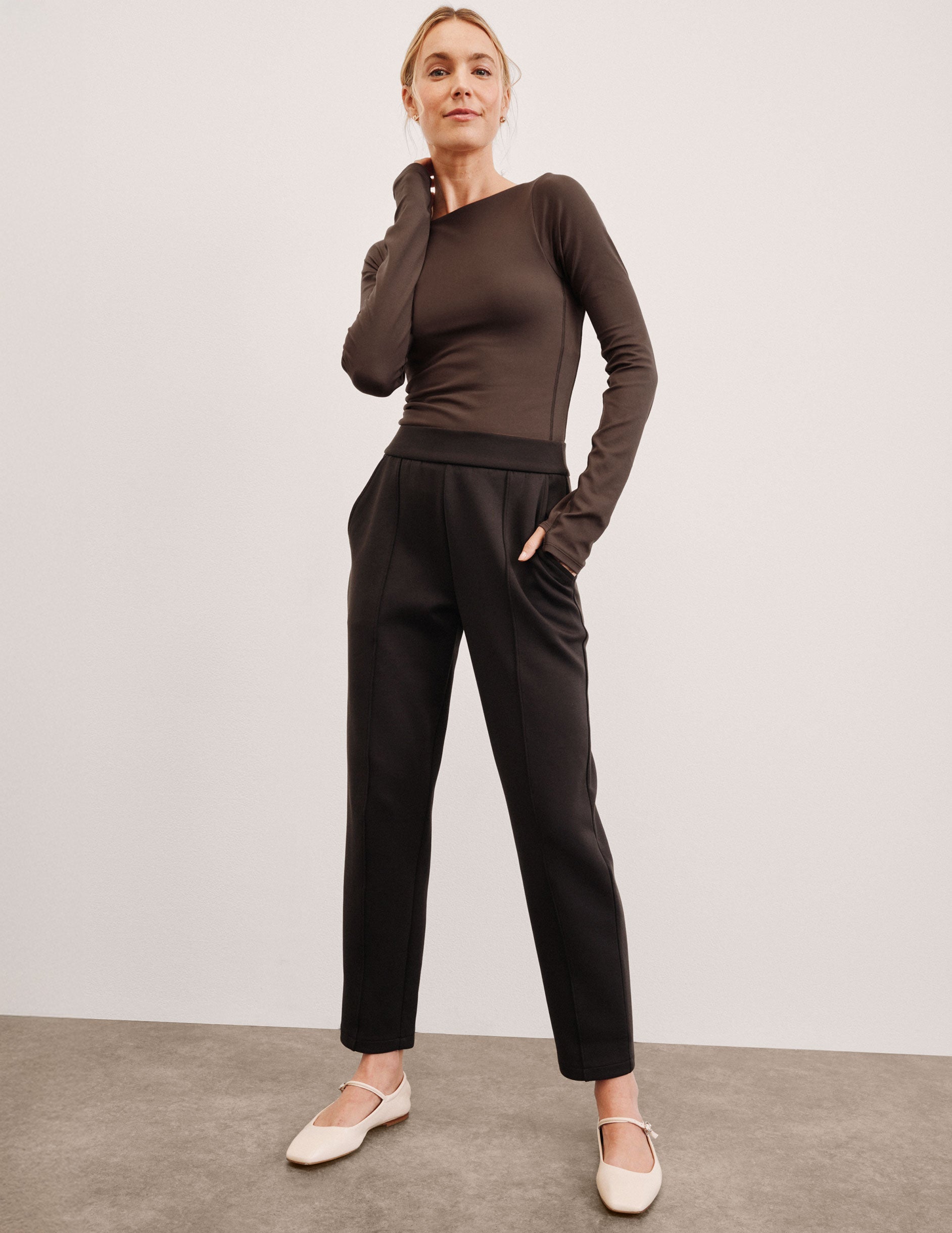 woman wearing a brown long sleeve and brown tapered pants against a plain background.