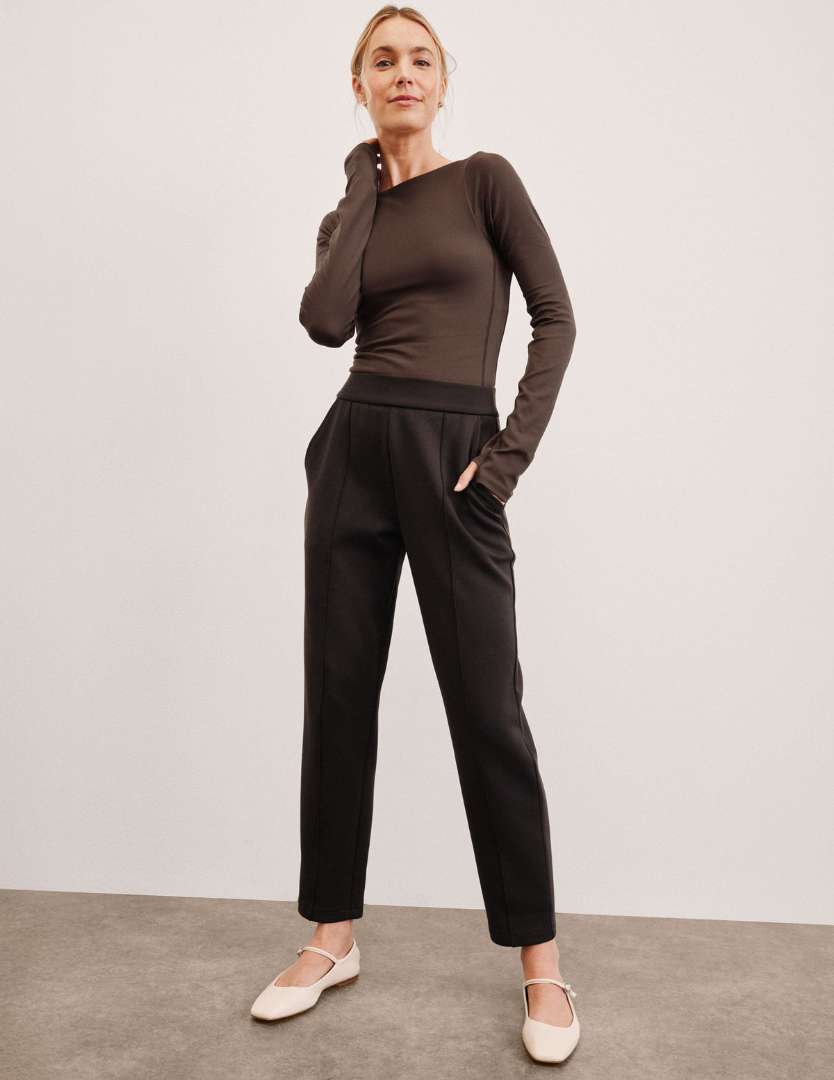 woman wearing a brown long sleeve and brown tapered pants against a plain background.