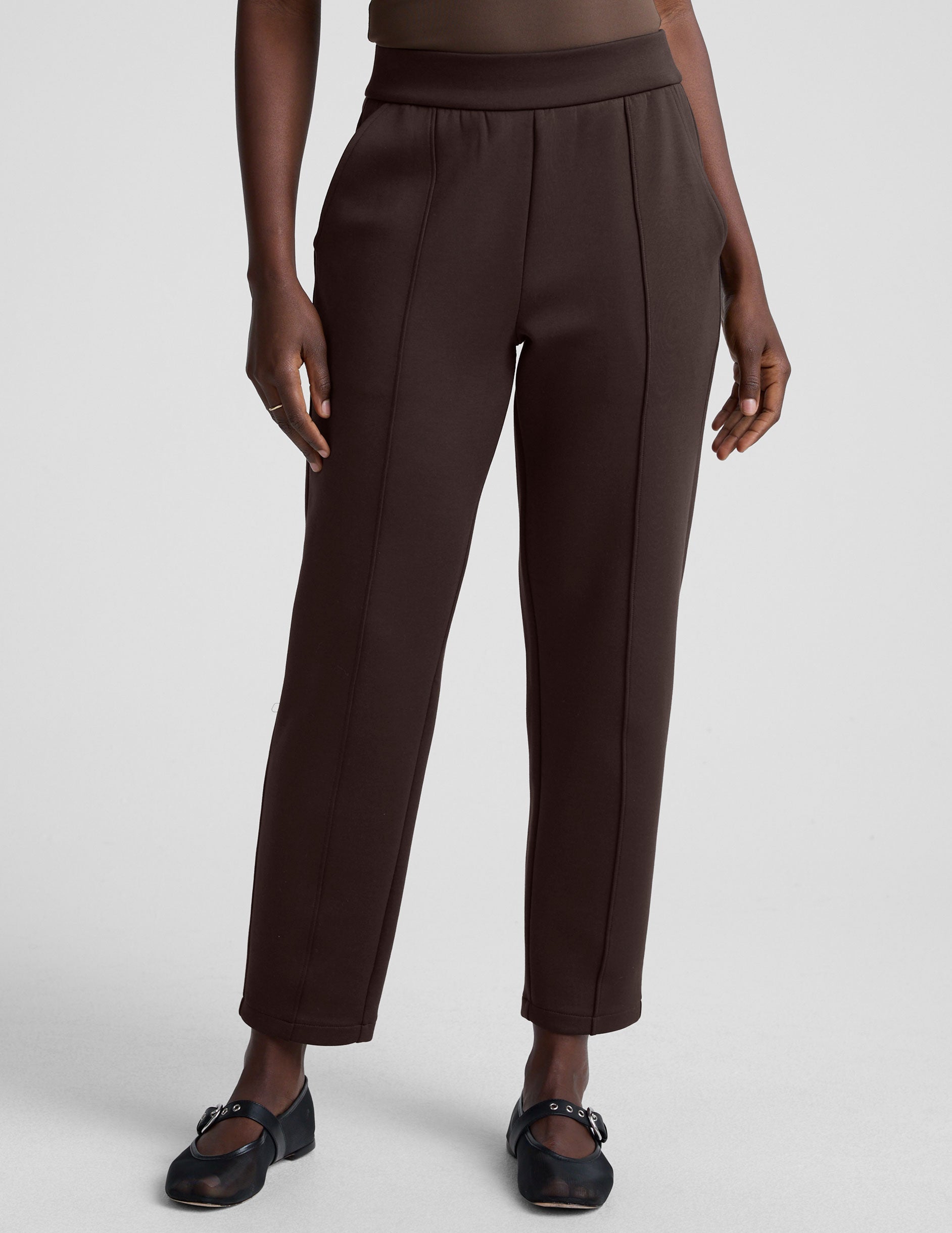 woman wearing brown tapered pants against a plain background.
