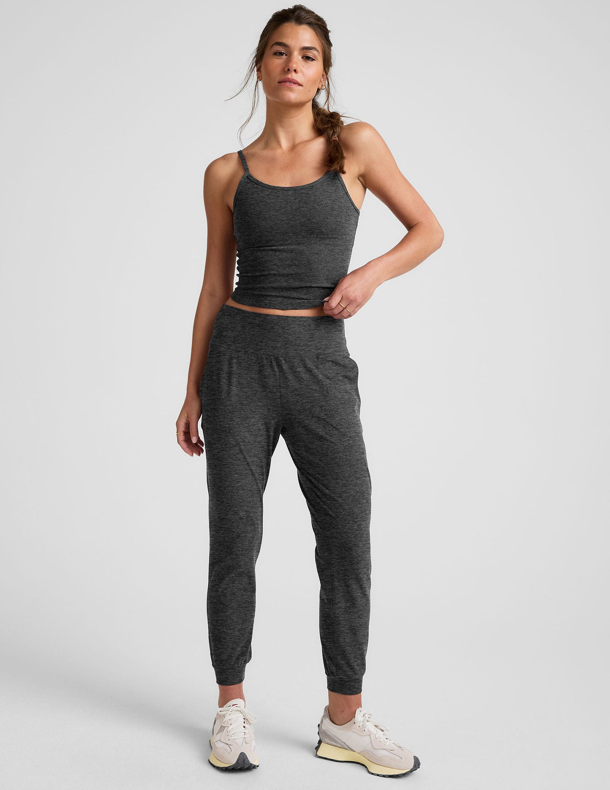woman wearing a dark gray tank top and joggers on a plain background.