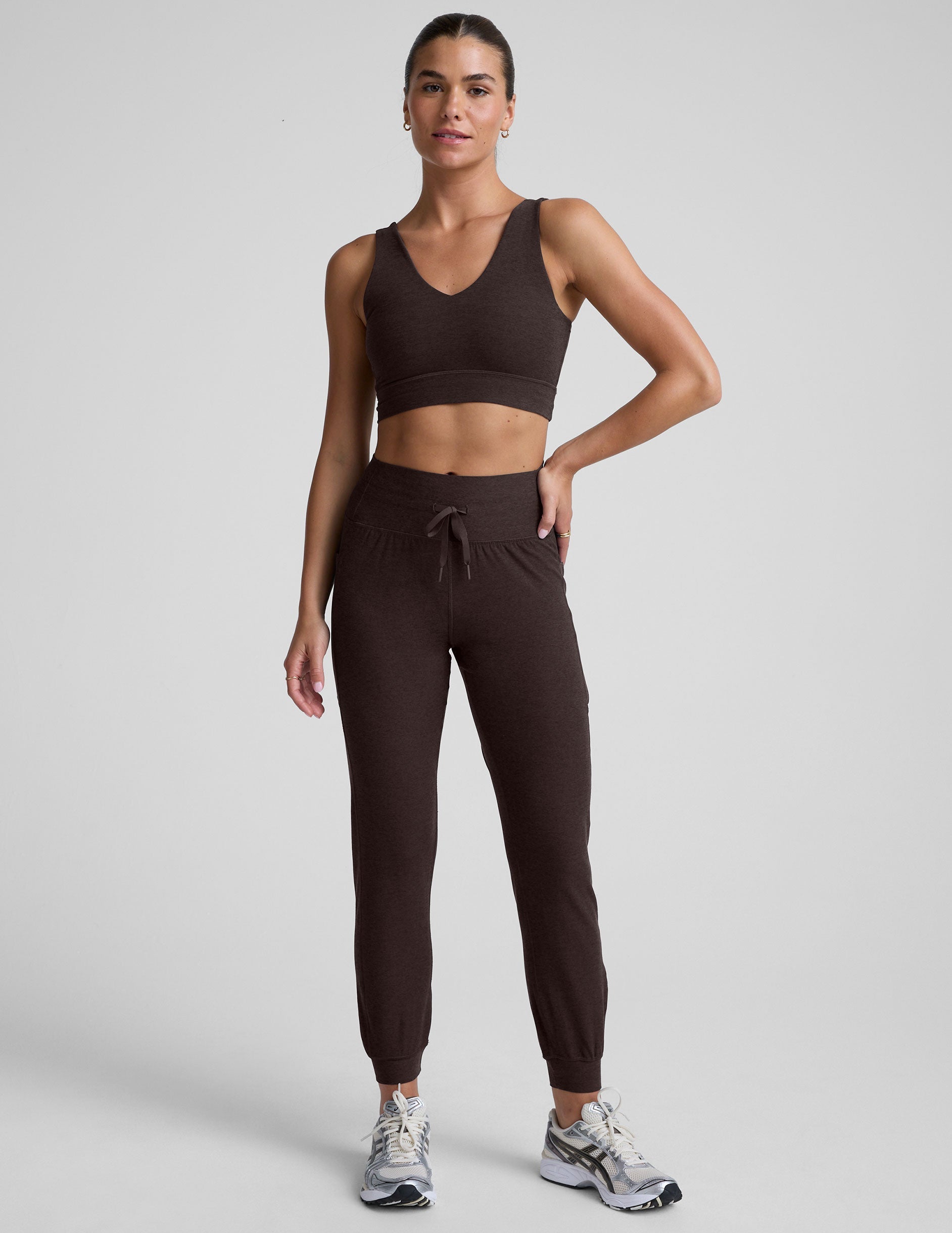 woman wearing a brown bra and joggers with pockets on a light gray background.
