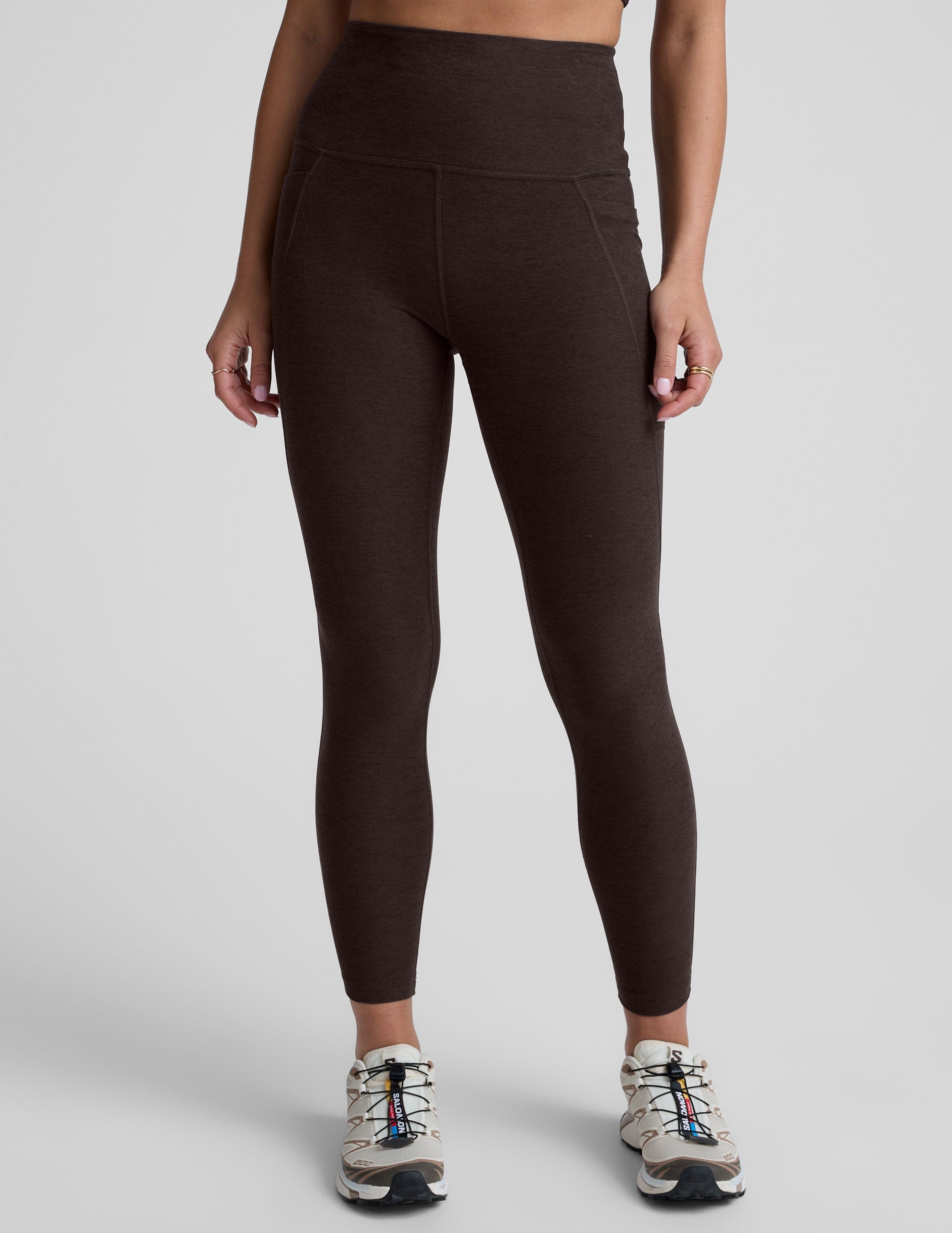 woman wearing a brown high-waisted midi length pocket leggings  with a plain background.