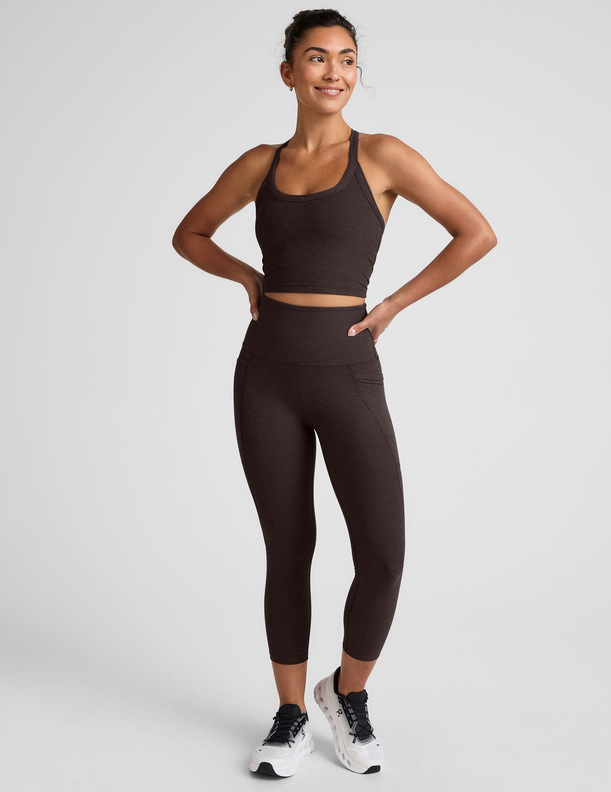 woman wearing a brown cropped tank and capri length leggings with pockets on a plain background.
