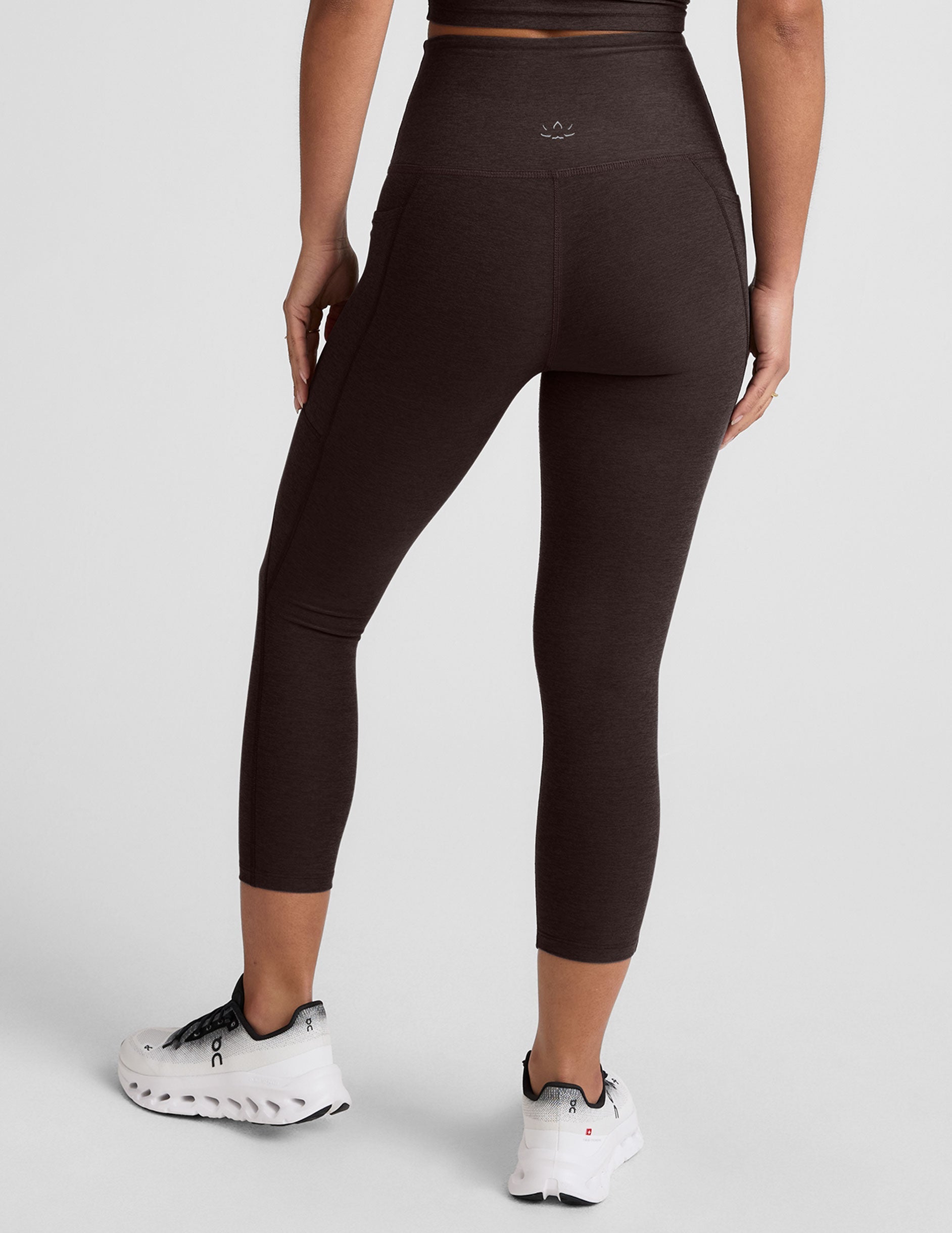 woman wearing a brown capri length leggings with pockets on a plain background.
