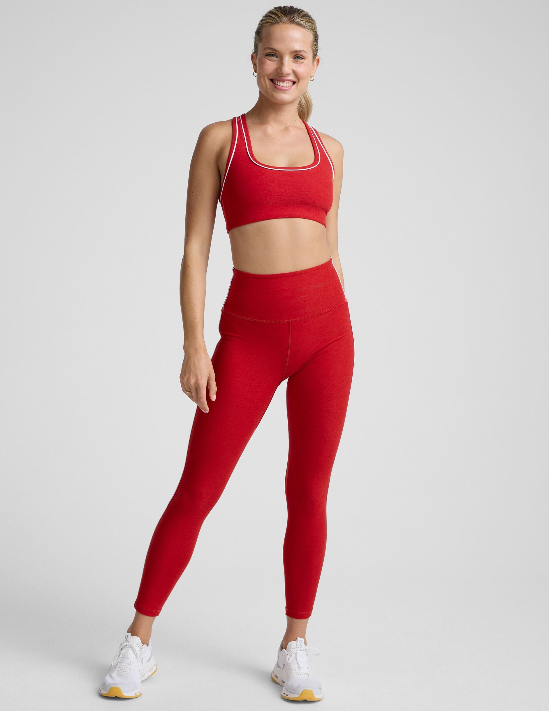 woman in red bra with white piping and red high-waisted midi length leggings with white piping against a plain background.