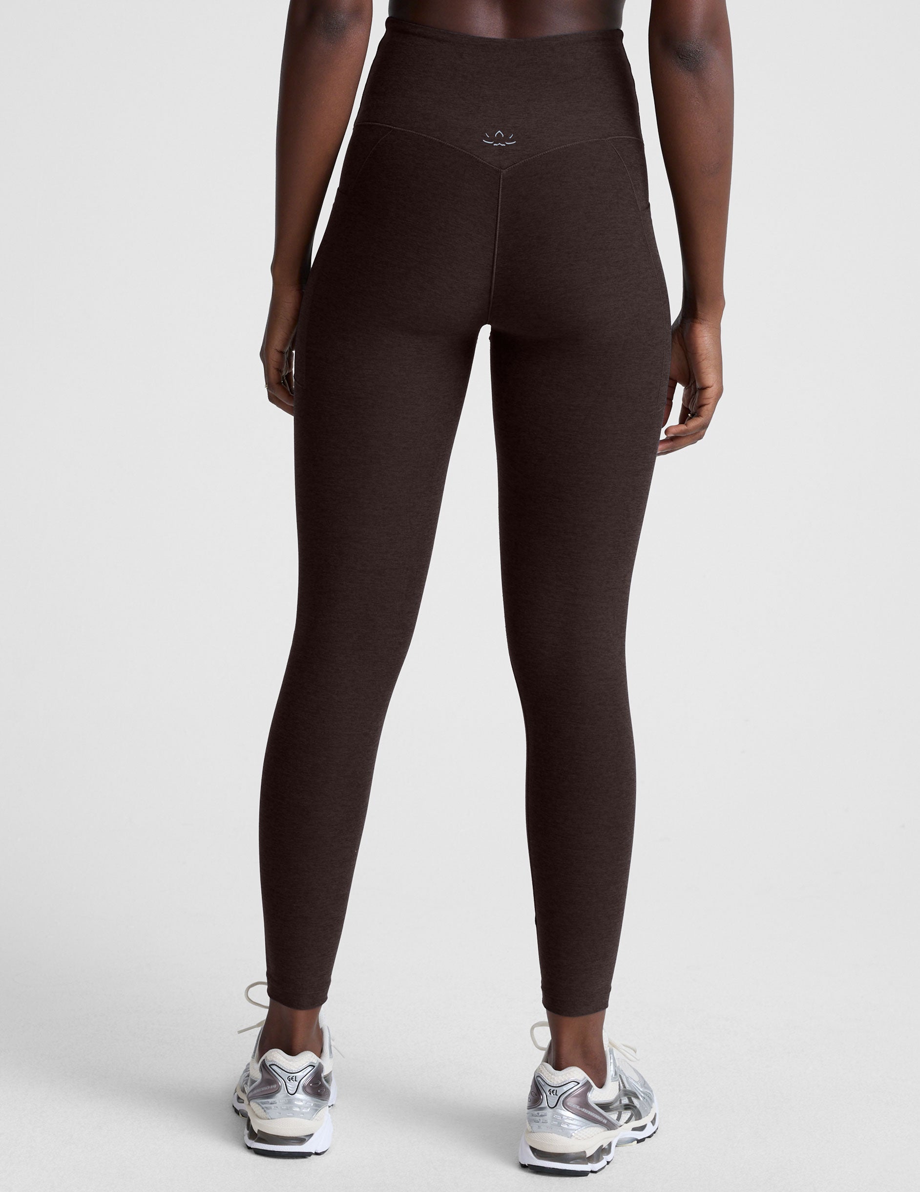 woman wearing brown high-waisted midi length leggings with pockets on a plain background.