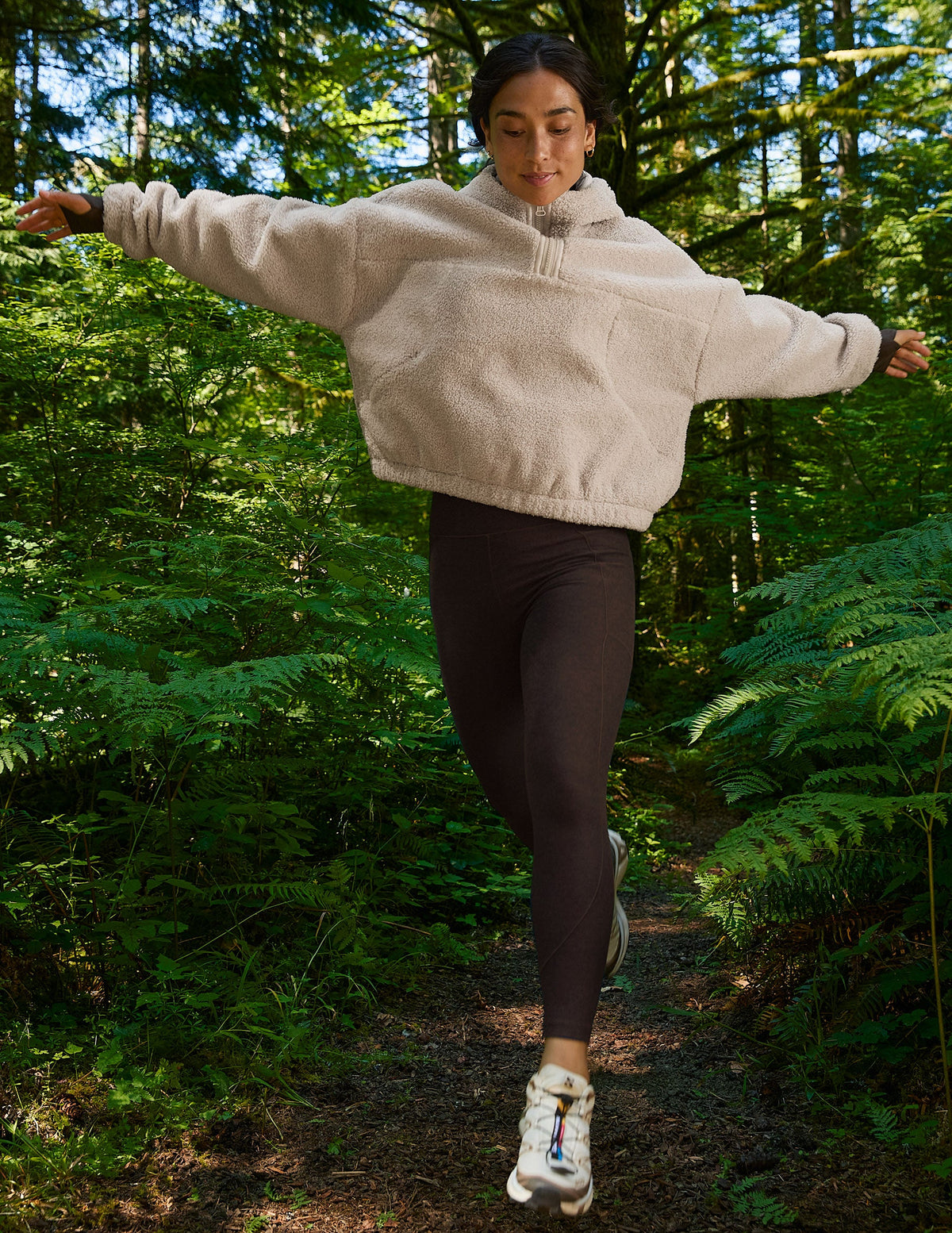 woman in a beige hoodie and brown pocket leggings running through a forest.