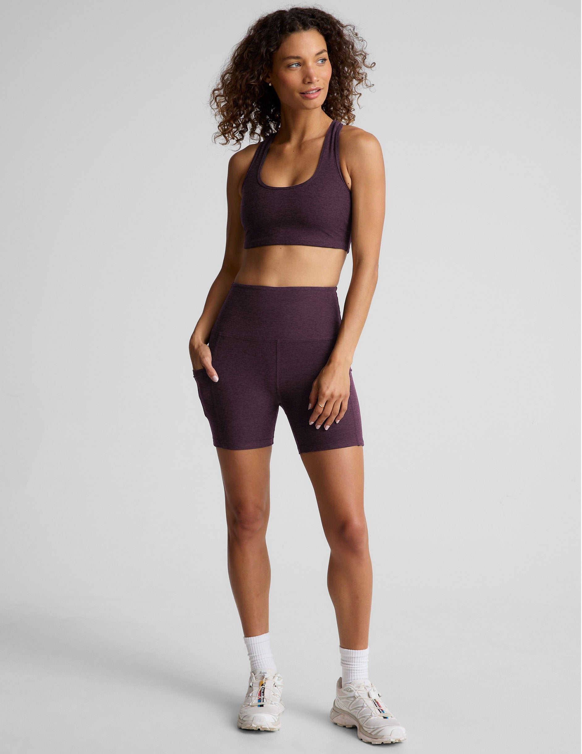model is wearing a purple sports bra and purple 5" biker shorts with pockets against a plain background. 