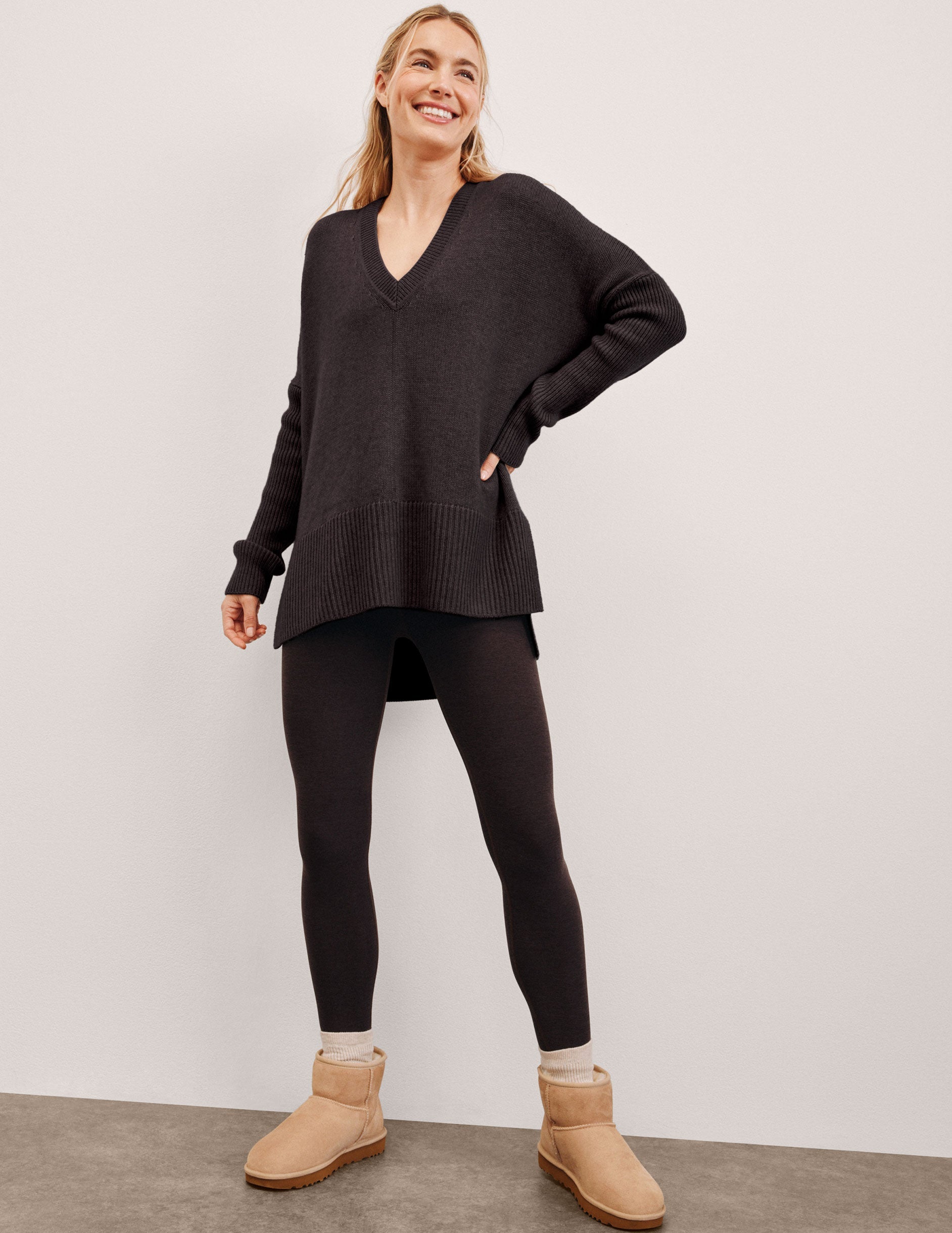 Woman wearing a brown sweater, black leggings, and beige boots against a plain background. 
